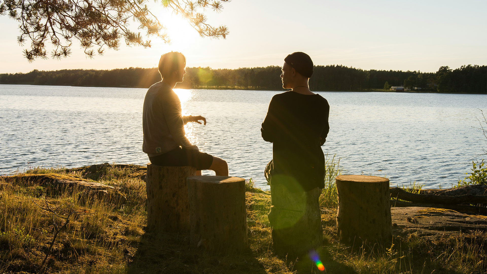 Zwei Männer sitzen bei Sonnenuntergang an einem See und unterhalten sich.